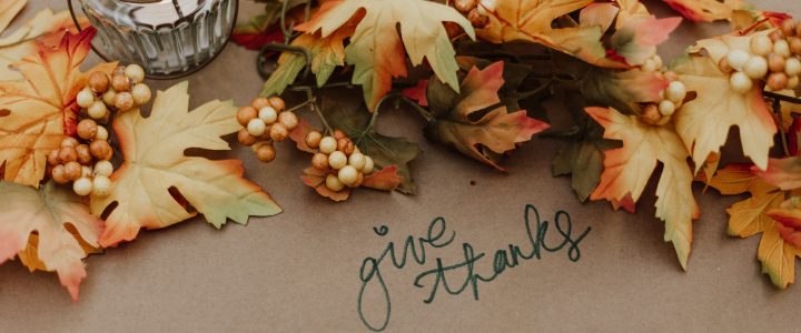 Fall leaves on a table with berries and a glass lantern. Handwritten text, give thanks.