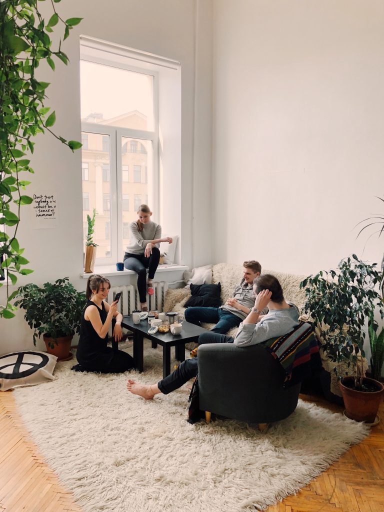2 men and 2 women sitting around a coffee table in their apartment, hanging out. White carpet, black chair, white patterned couch, and plants. 