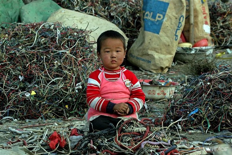 Small child in red jumper sitting amidst a pile of wires in Guiyu, China at an e-waste salvage area. 