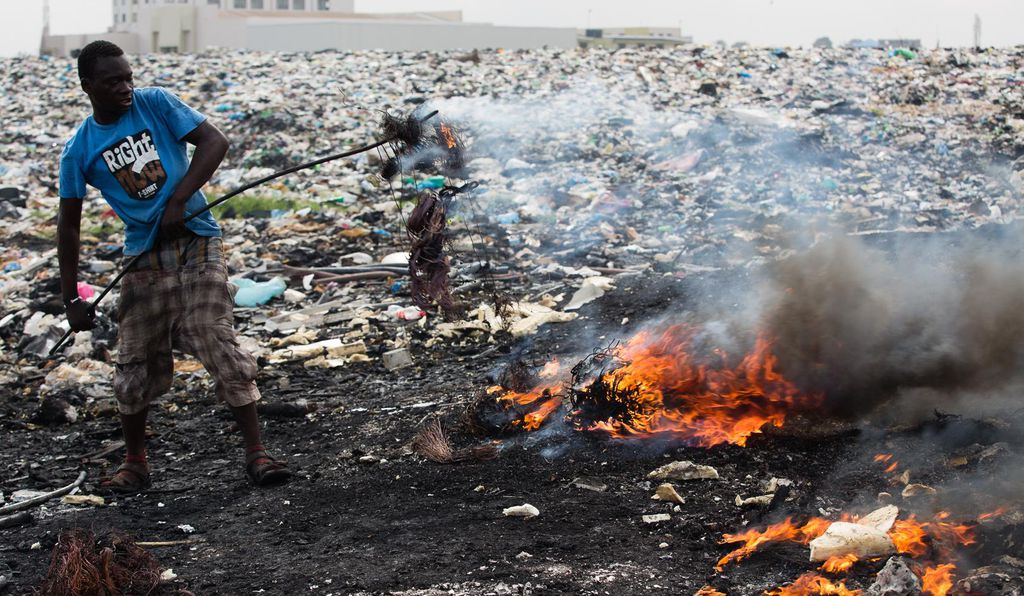 Man in blue t-shirt and shorts burning e-waste at a dump site in Agbogboshie, Ghana. 