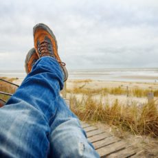 Person wearing blue denim jeans and hiking boots, only showing their legs as they look at a beach