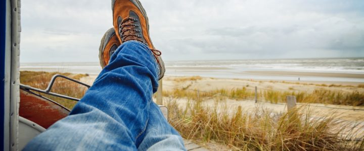 Person wearing blue denim jeans and hiking boots, only showing their legs as they look at a beach
