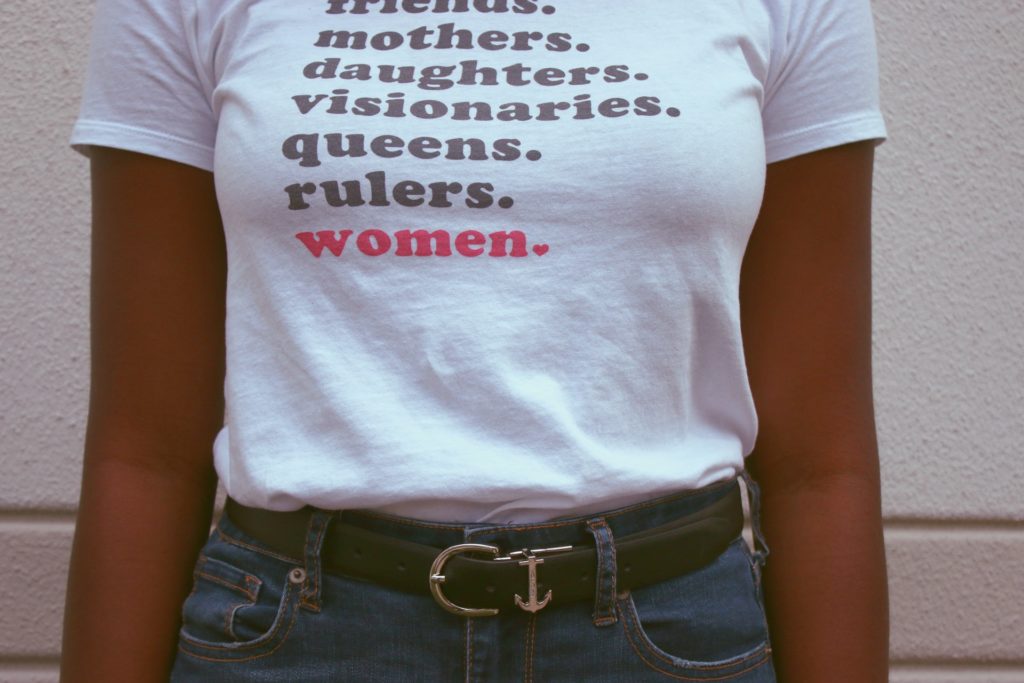 Black woman wearing white short sleeved t-shirt with woman focused words written on it. Cropped with her head not showing.