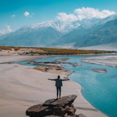 Man standing on a rock on a sandy bank beside a river in front of snow capped mountains