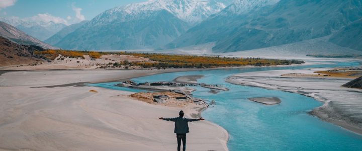 Man standing on a rock on a sandy bank beside a river in front of snow capped mountains