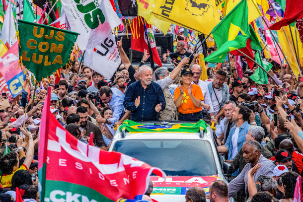 President-Elect Lula standing on the back of a pick up truck on the campaign trail pre election, with wife Janja by his side. Throngs of adoring fans wave flags and try to get close to him.