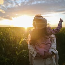 Woman holding a small child in a field of green at sunset. Child pointing toward the sky.
