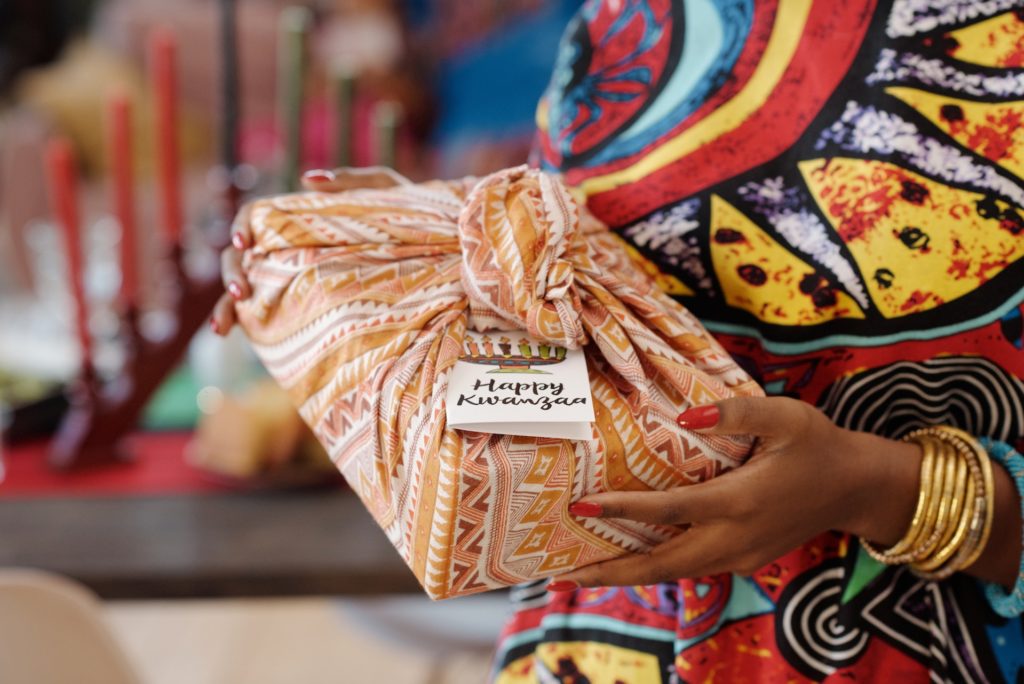 Woman in traditional African clothing giving a Kwanzaa gift wrapped in decorative orange and brown colored fabric.