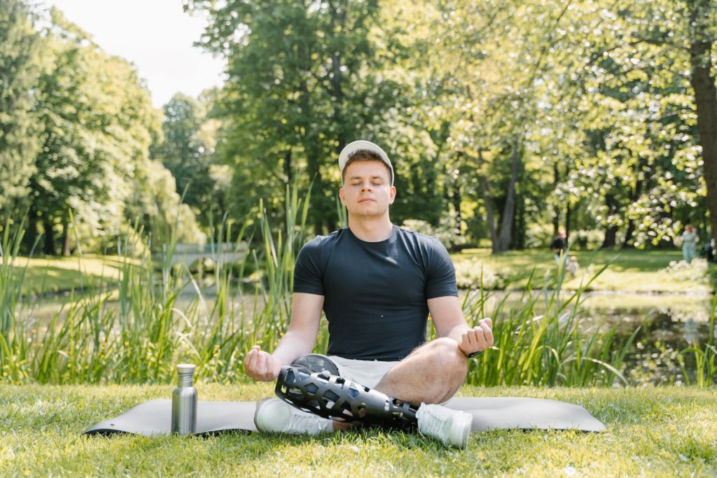 White man with one prosthetic leg sitting cross legged on a yoga mat, in a zen pose, in a park with long green grass and trees behind him.