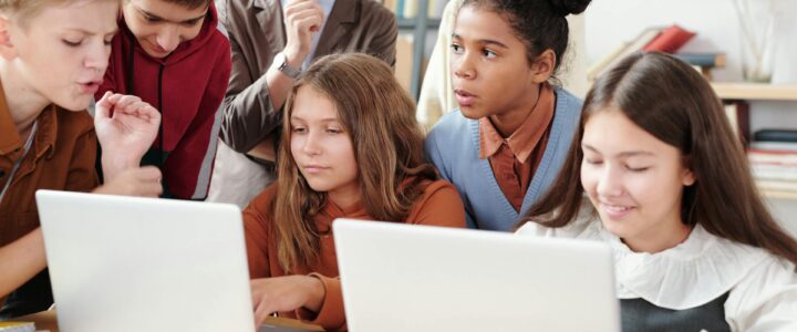 Group of students looking at laptops with teacher in the background