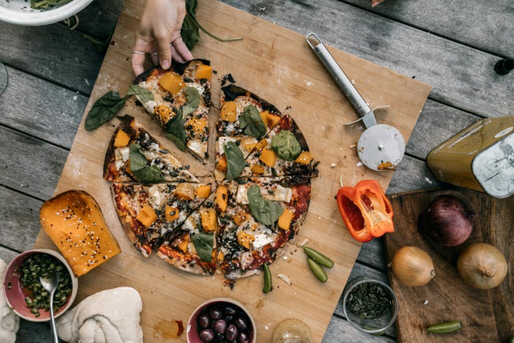 Vegetarian pizza with pumpkin, red pepper, red onion, brown onion, baby spinach, pepitas and capers. Being served on a wooden chopping board, sliced up.